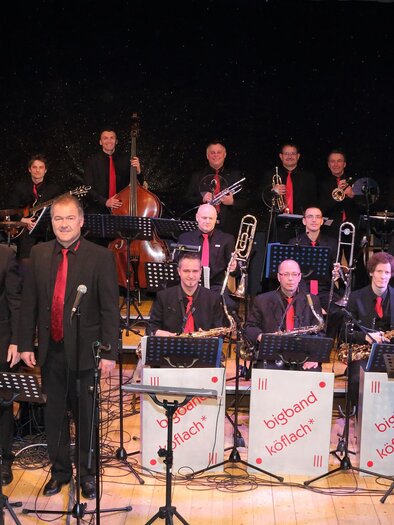 Eine große Gruppe von Musikern steht auf der Bühne, die in formeller Kleidung gekleidet sind. Im Vordergrund stehen einige Sänger, während die Band im Hintergrund spielt. | © Bigband Köflach