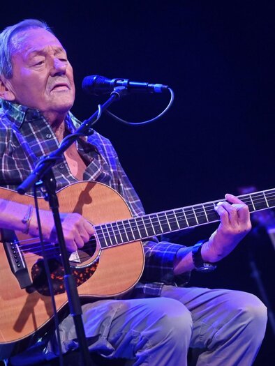 Wolfgang Ambros auf der Bühne mit Gitarre in Händen auf einem Hocker sitzend, davor das Mikrofon in einem blauen Bühnenlicht.  | © Dietmar Lipkovich