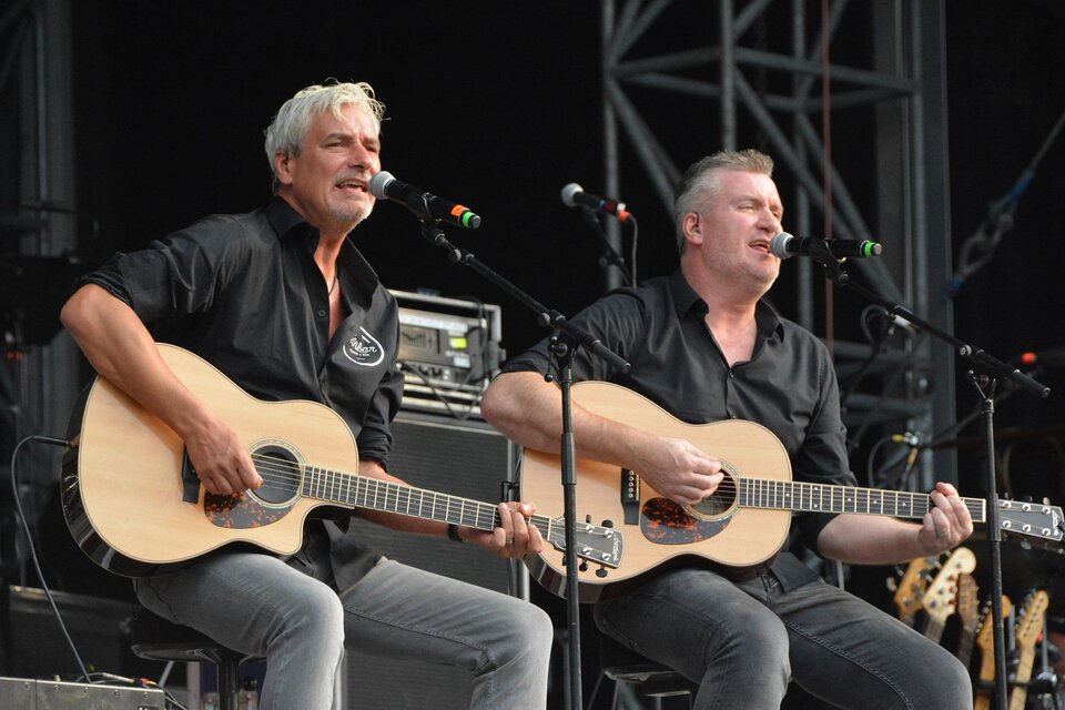 Zwei Männer sitzen auf einer Bühne und spielen akustische Gitarren. Sie singen in ein Mikrofon und scheinen ein Konzert zu geben. | © de Zwa