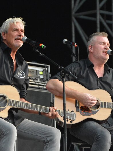 Zwei Männer sitzen auf einer Bühne und spielen akustische Gitarren. Sie singen in ein Mikrofon und scheinen ein Konzert zu geben. | © de Zwa