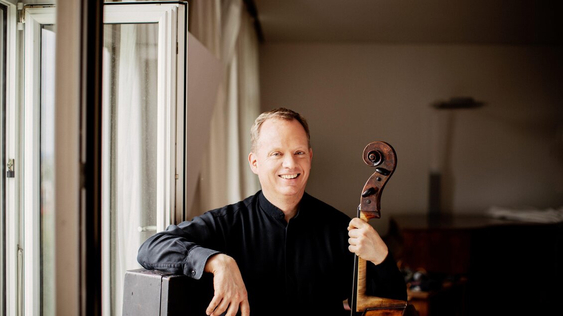Cellist Christoph Stradner steht mit seinem Violoncello neben einem geöffneten Fenster. | © Julia Wesely
