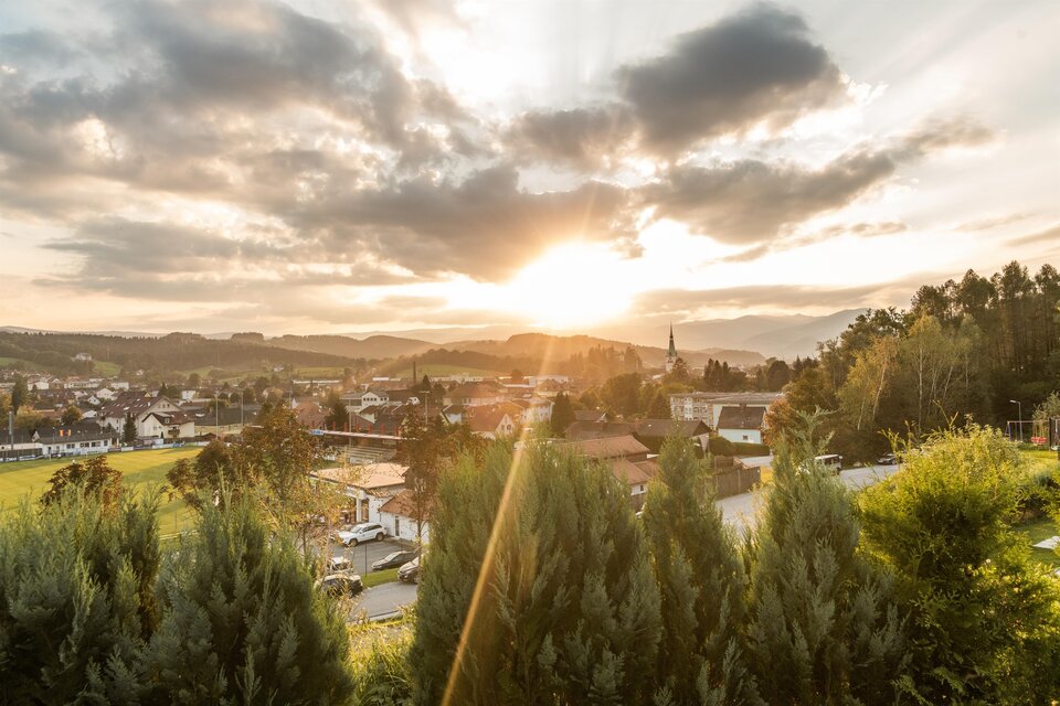 Eine malerische Landschaft mit einem Sonnenuntergang über einer Stadt. Die Bäume im Vordergrund und die Berge im Hintergrund schaffen eine ruhige Atmosphäre. | © Lipizzanerheimat-Die Abbilderei