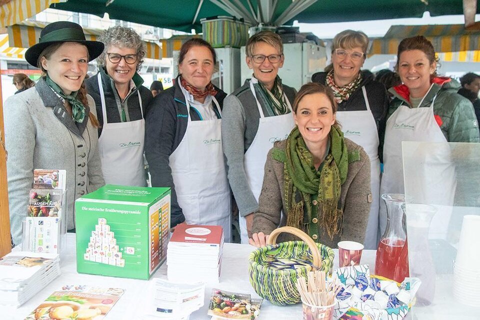 Eine Gruppe von sieben freundlichen Frauen steht an einem Marktstand. Sie tragen Schürzen und lächeln in die Kamera, während verschiedene Lebensmittel und Produkte auf dem Tisch präsentiert werden. | © Pressberger