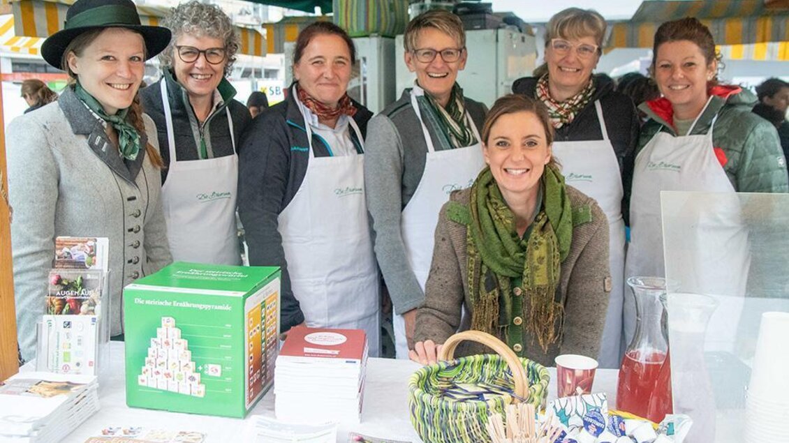Eine Gruppe von sieben freundlichen Frauen steht an einem Marktstand. Sie tragen Schürzen und lächeln in die Kamera, während verschiedene Lebensmittel und Produkte auf dem Tisch präsentiert werden. | © Pressberger