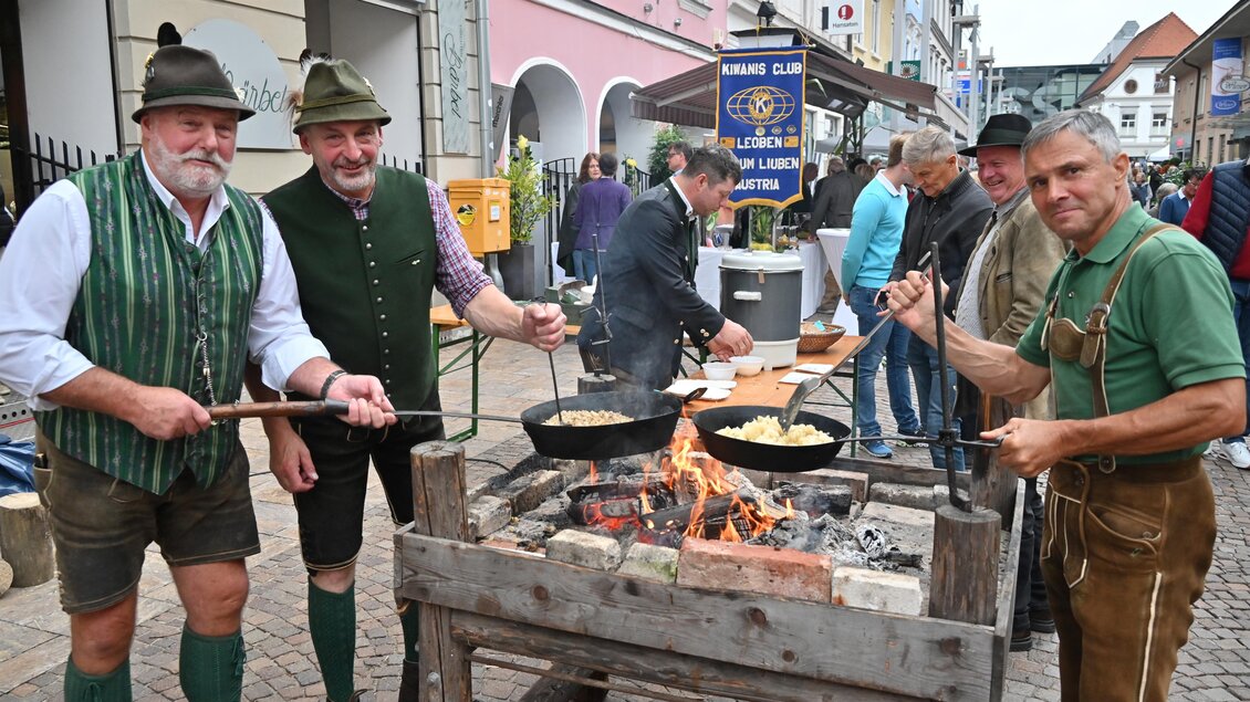 Eine Gruppe von Männern in traditioneller Tracht steht um ein Feuer und kocht in großen Pfannen. Im Hintergrund sind Straßen und andere Personen zu sehen. | © Norbert Ortner