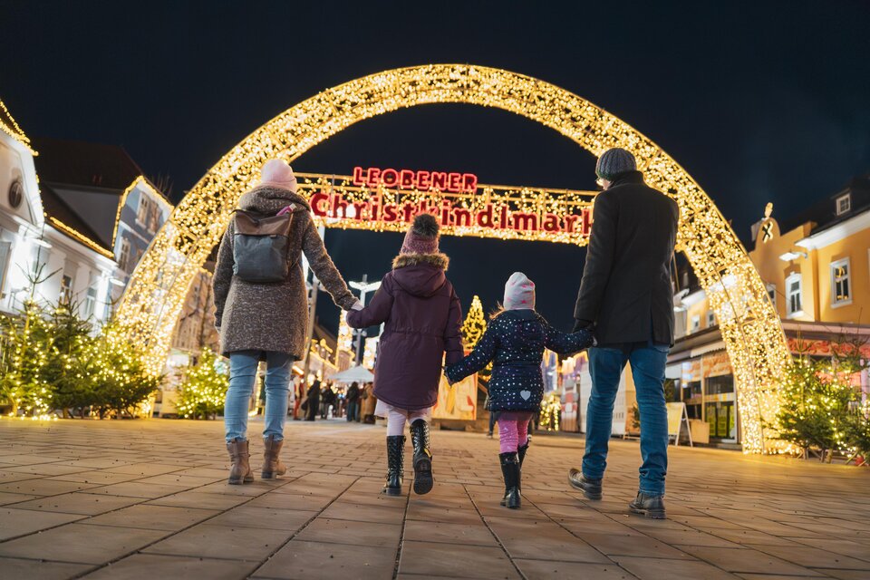Eine Familie spaziert unter einem festlich beleuchteten Bogen in einem Weihnachtsmarkt. Die Szenerie ist abends und voller Lichter, die eine gemütliche Stimmung erzeugen. | © TVB Erzberg-Leoben