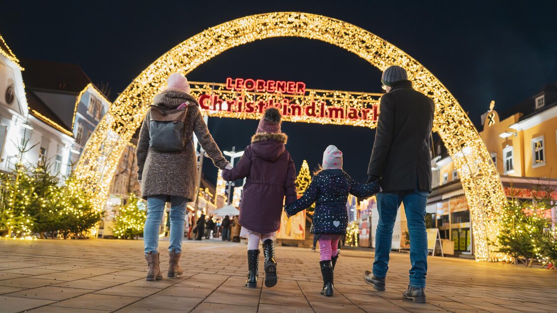Eine Familie spaziert unter einem festlich beleuchteten Bogen in einem Weihnachtsmarkt. Die Szenerie ist abends und voller Lichter, die eine gemütliche Stimmung erzeugen. | © TVB Erzberg-Leoben