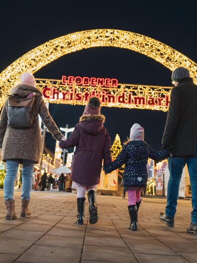 A family walks under a festively lit arch at a Christmas market. The scene is in the evening and full of lights, creating a cozy atmosphere. | © TVB Erzberg-Leoben