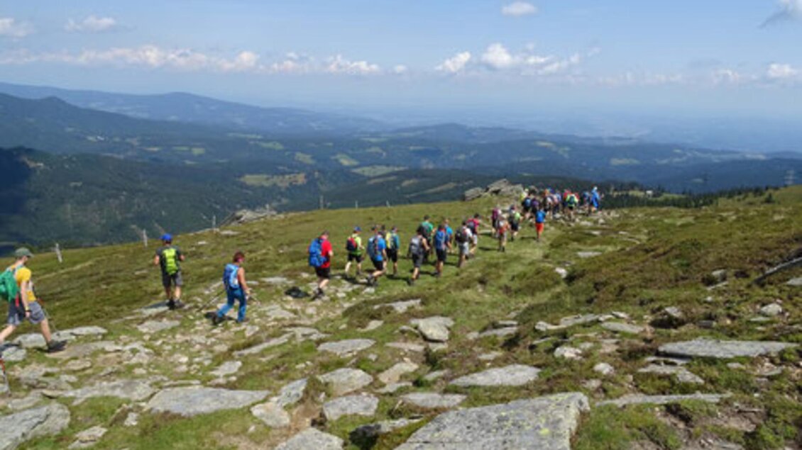 Eine Gruppe von Wanderern bewegt sich über einen felsigen Pfad in einer hügeligen Landschaft. Der Himmel ist blau mit einigen Wolken und die Aussicht ist beeindruckend. | © Koralm24