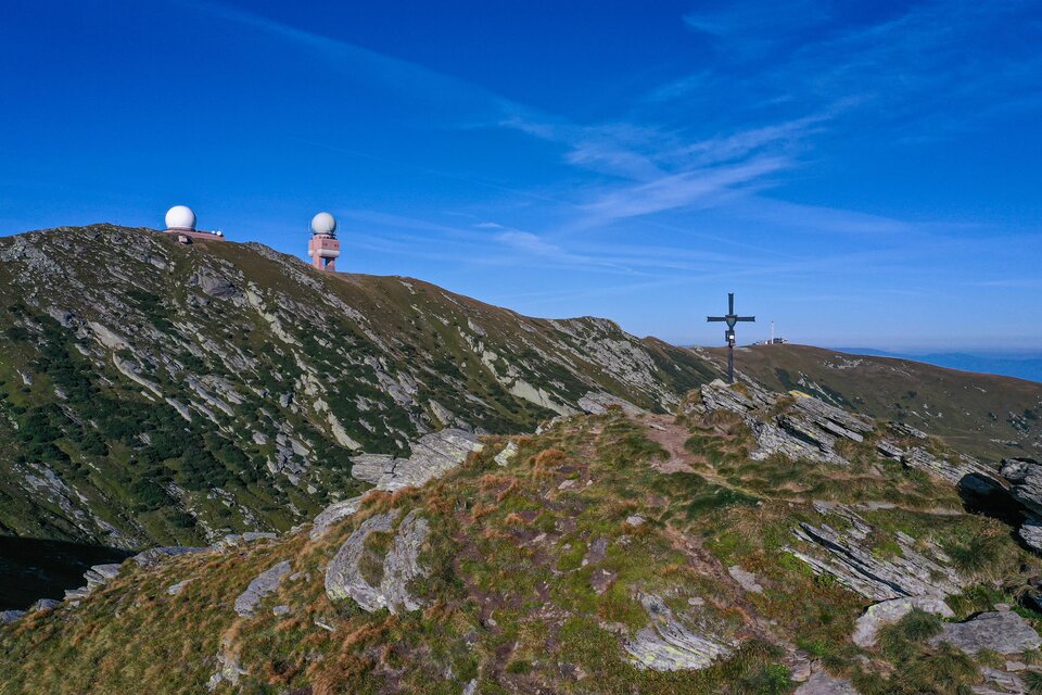 Eine hügelige Landschaft mit zwei großen Radarstationen auf einem Gipfel. Im Vordergrund steht ein Kreuz auf einem felsigen Gelände. | © Christian Freydl
