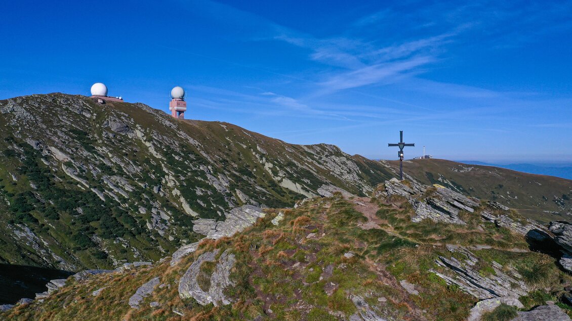Eine hügelige Landschaft mit zwei großen Radarstationen auf einem Gipfel. Im Vordergrund steht ein Kreuz auf einem felsigen Gelände. | © Christian Freydl