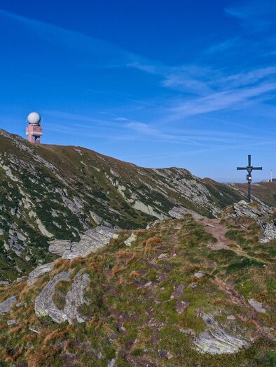 Eine hügelige Landschaft mit zwei großen Radarstationen auf einem Gipfel. Im Vordergrund steht ein Kreuz auf einem felsigen Gelände. | © Christian Freydl