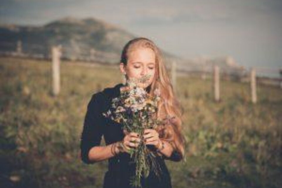 Ein Mädchen mit langen blonden Haaren steht auf einer Wiese und hält einen Strauß Blumen. Im Hintergrund sind sanfte Hügel sichtbar. | © nicoleseiser.at