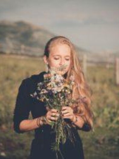 Ein Mädchen mit langen blonden Haaren steht auf einer Wiese und hält einen Strauß Blumen. Im Hintergrund sind sanfte Hügel sichtbar. | © nicoleseiser.at