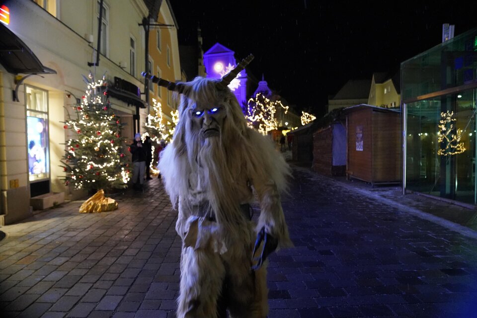 Ein mysteriöser, händlerähnlicher Charakter mit Hörnern steht auf einer festlich beleuchteten Straße. Im Hintergrund sind Weihnachtsbäume und dekorative Lichter sichtbar. | © Stadtgemeinde Frohnleiten