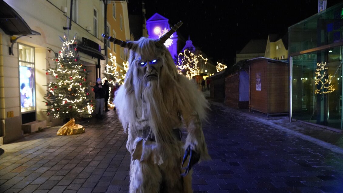 Ein mysteriöser, händlerähnlicher Charakter mit Hörnern steht auf einer festlich beleuchteten Straße. Im Hintergrund sind Weihnachtsbäume und dekorative Lichter sichtbar. | © Stadtgemeinde Frohnleiten