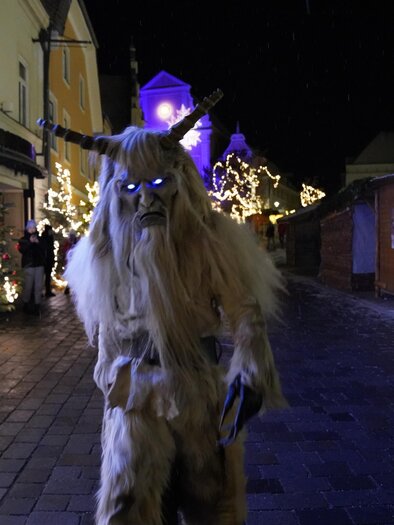 Ein mysteriöser, händlerähnlicher Charakter mit Hörnern steht auf einer festlich beleuchteten Straße. Im Hintergrund sind Weihnachtsbäume und dekorative Lichter sichtbar. | © Stadtgemeinde Frohnleiten