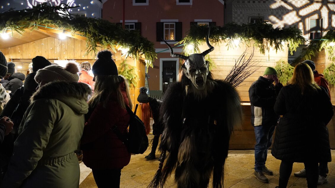 Ein festlicher Weihnachtsmarkt mit Besuchern und einem skurrilen Maskenträger im Vordergrund. Die Stimmung ist lebhaft und gemütlich, umgeben von Lichtern und Dekorationen. | © Stadtgemeinde Frohnleiten