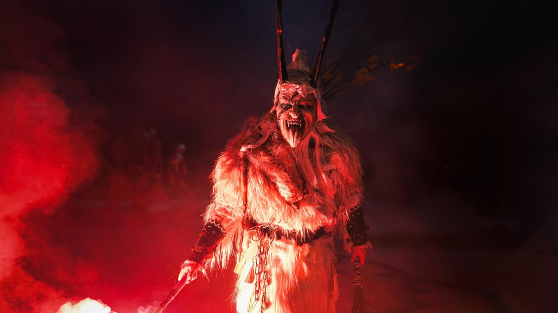 Ein mystischer Charakter in einer Fellkostüm mit einer Maske steht im roten Licht. Der Hintergrund ist dunkel und rauchig, was eine spannende Atmosphäre schafft. | © Stadtgemeinde Bärnbach