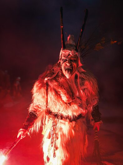 Ein mystischer Charakter in einer Fellkostüm mit einer Maske steht im roten Licht. Der Hintergrund ist dunkel und rauchig, was eine spannende Atmosphäre schafft. | © Stadtgemeinde Bärnbach