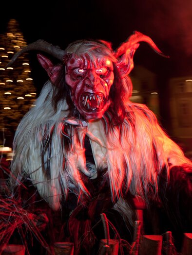 Krampus mit Hörnern, langen Haaren und furchterregender Maske, rot beleuchtet beim nächtlichen Krampuslauf in Voitsberg. | © Graz Tourismus-Harry Schiffer