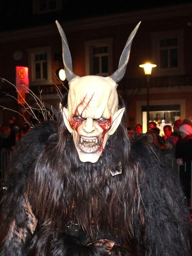 A spooky, masked character with horns and a terrifying grimace. In the background, spectators can be seen at a nighttime parade. | © TV Südsteiermark - Irene Löschnig
