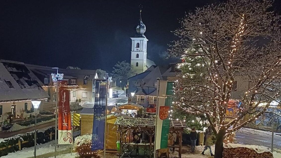 Eine verschneite Stadt mit festlich beleuchtetem Weihnachtsbaum und einer Kirche im Hintergrund. Die Atmosphäre ist ruhig und festlich in der Nacht. | © Gemeinde St. Stefan