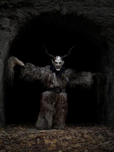 A creepy figure with a skull face and horned head emerges from a dark cave. The ground is covered with leaves and the atmosphere is gloomy.