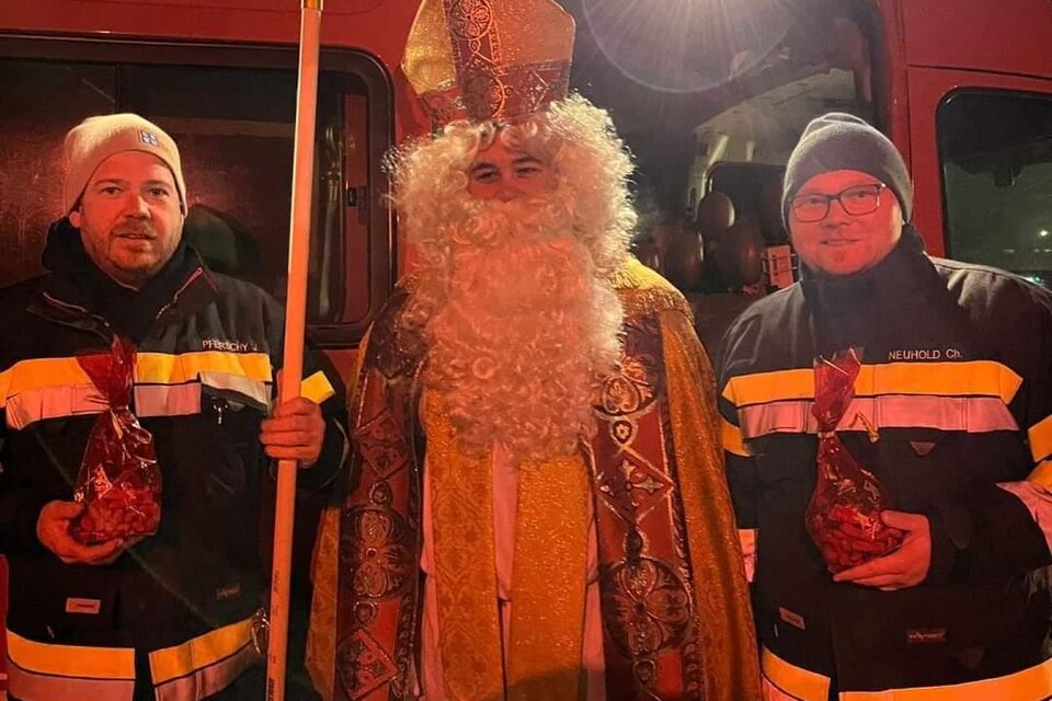 Eine festliche Szene mit einem Mann in einem roten Gewand und mit einem Bischofsstab, umgeben von zwei Personen in Feuerwehrkleidung. Im Hintergrund ist ein Feuerwehrfahrzeug zu sehen. | © FF Bad Waltersdorf