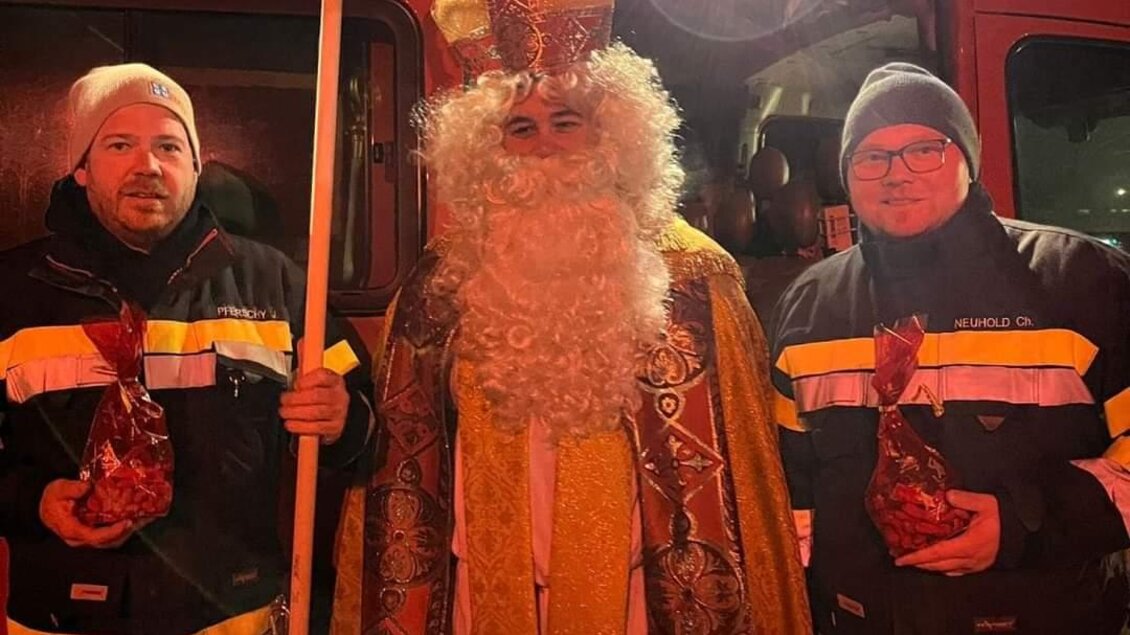 Eine festliche Szene mit einem Mann in einem roten Gewand und mit einem Bischofsstab, umgeben von zwei Personen in Feuerwehrkleidung. Im Hintergrund ist ein Feuerwehrfahrzeug zu sehen. | © FF Bad Waltersdorf