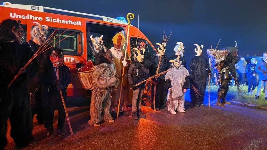 Eine Gruppe von Menschen in traditionellen Kostümen und Masken steht auf einer Straße. Im Hintergrund ist ein Einsatzfahrzeug mit der Aufschrift „Unsre Sicherheit“ zu sehen. | © FF Bad Waltersdorf