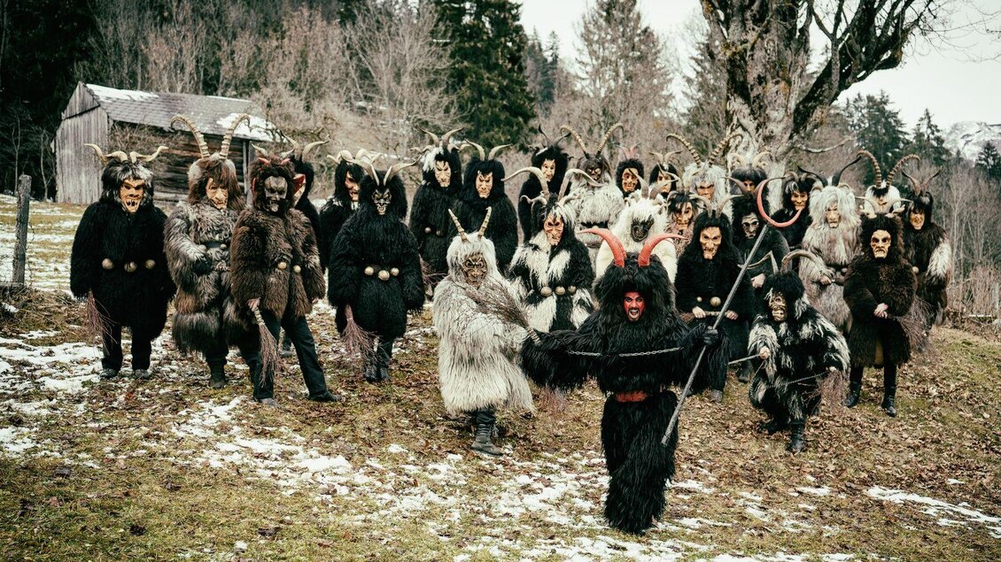 Luzifer mit rot geschminktem Gesicht steht gemeinsam mit einer Gruppe von Krampussen in hellen und dunklen Mänteln sowie schaurigen Krampusmasken. Er hält seinen Luziferstab in die Höhe und wird von seinen beiden Haltern an Ketten festgehalten. | © Lebensbilder - Christine Struz