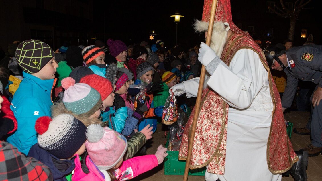 Der Nikolaus kniet vor den aufgeregten Kindern und verteilt seine Nikolosackerln. Die Kinder tragen bunte Winterkleidung und blicken gespannt zu ihm auf. In seiner linken Hand hält der Nikolaus seinen Stab. Er ist festlich in Weiß und Rot gekleidet, wobei sein langer, weißer Bart und die weißen Handschuhe besonders hervorstechen. | © Lebensbilder - Christine Struz