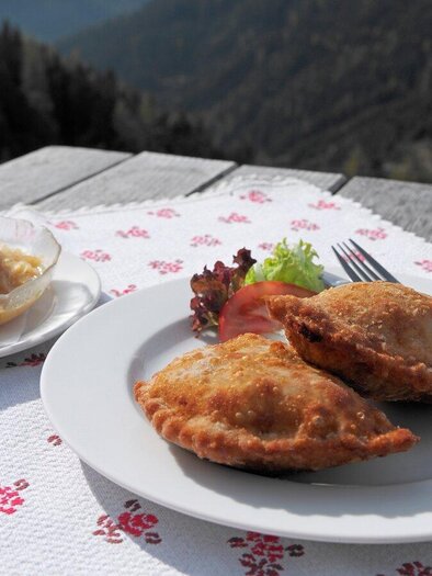 Ein Teller mit frittierten Teigtaschen und einer Schale mit Beilagen auf einem Holztisch. Im Hintergrund sind Berglandschaften zu sehen. | © Herbert Raffalt