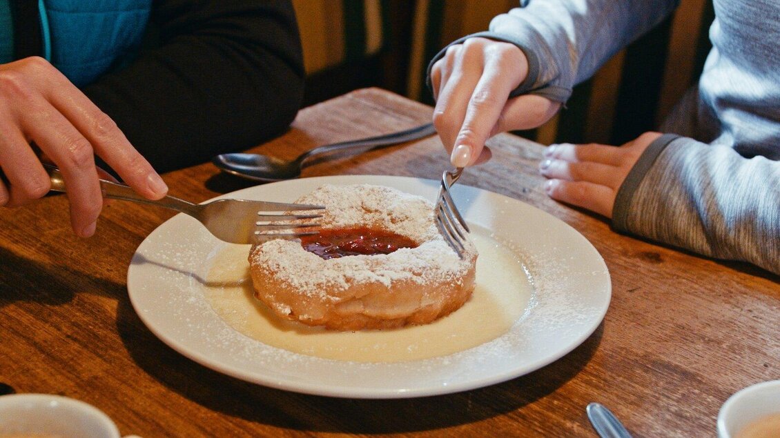 Zwei Hände schneiden gemeinsam einen süßen Kuchen auf einem Teller. Der Kuchen ist mit Puderzucker bestreut und hat eine fruchtige Füllung. | © Gerald Grünwald