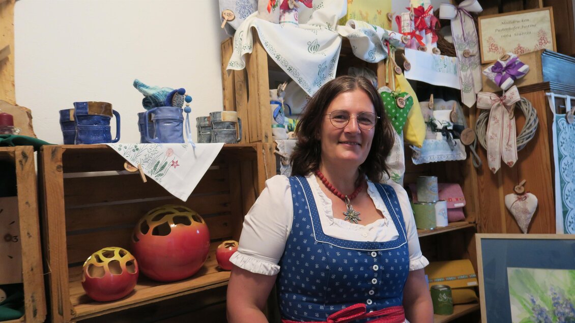 Eine Frau in traditioneller Tracht steht vor einem dekorierten Holzregal. Im Hintergrund sind handgefertigte Gegenstände und Dekorationen zu sehen. | © Marlene Zandl