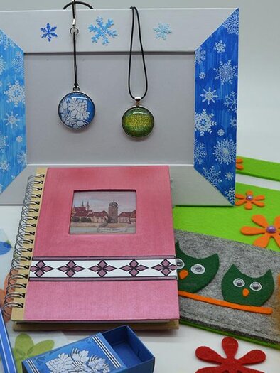 Colorful crafting materials and a notebook are placed on a table. In the background, two creative pendants are hanging in a decorative frame. | ©  MuseumsCenter