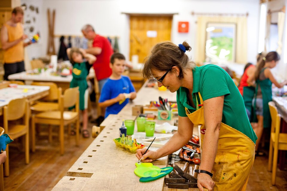 Ein kreativer Workshop mit Kindern, die an verschiedenen Kunstprojekten arbeiten. Eine Frau mit einer Schürze zeichnet konzentriert an einem Tisch voller Bastelmaterialien. | © Holzmuseum