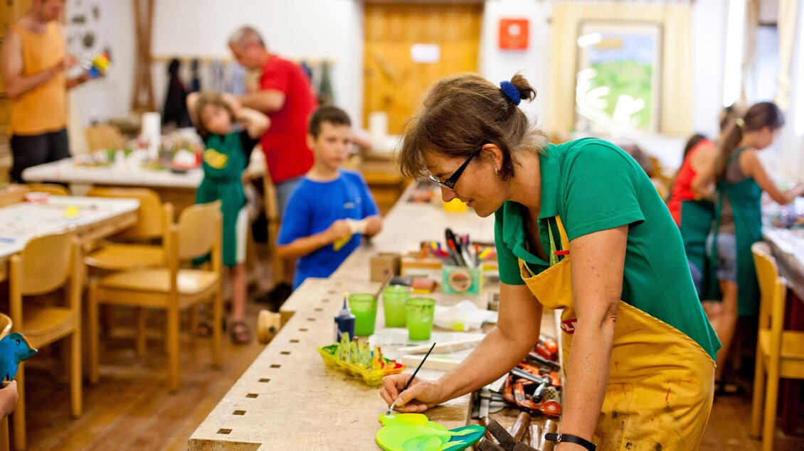 Ein kreativer Workshop mit Kindern, die an verschiedenen Kunstprojekten arbeiten. Eine Frau mit einer Schürze zeichnet konzentriert an einem Tisch voller Bastelmaterialien. | © Holzmuseum