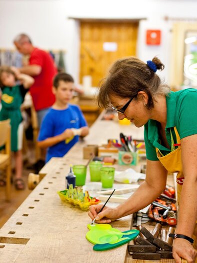 A creative workshop with children working on various art projects. A woman in an apron is intently drawing at a table full of crafting materials. | © Holzmuseum
