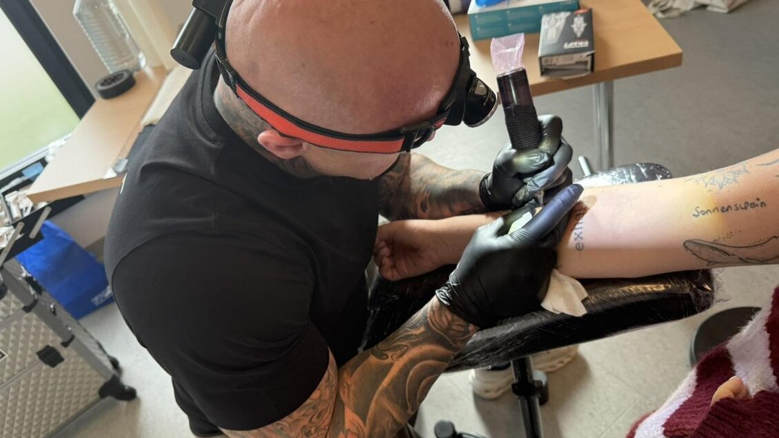 Ein Tätowierer arbeitet an einem Armtattoo. Es sind verschiedene Utensilien auf dem Tisch sichtbar. | © Pfleger