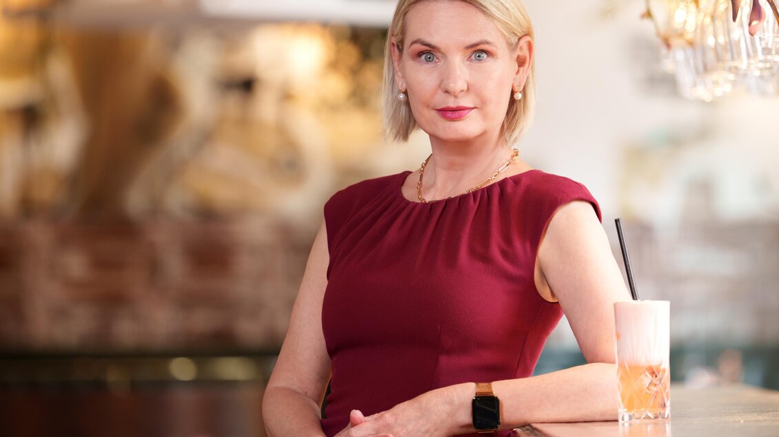 Claudia Rossbacher posiert bei einer Kommode mit einem Getränk daneben, Lesung im Römerhof Eggersdorf. | © Lucija Novak