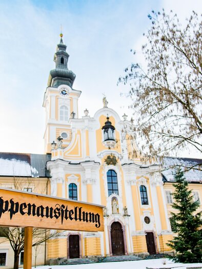 Eine beeindruckende Kirche mit einem hohen Turm und schöner Architektur. Vor der Kirche steht ein Schild mit der Aufschrift „Krippenausstellung“. | © TV Region Graz - Mias Photoart