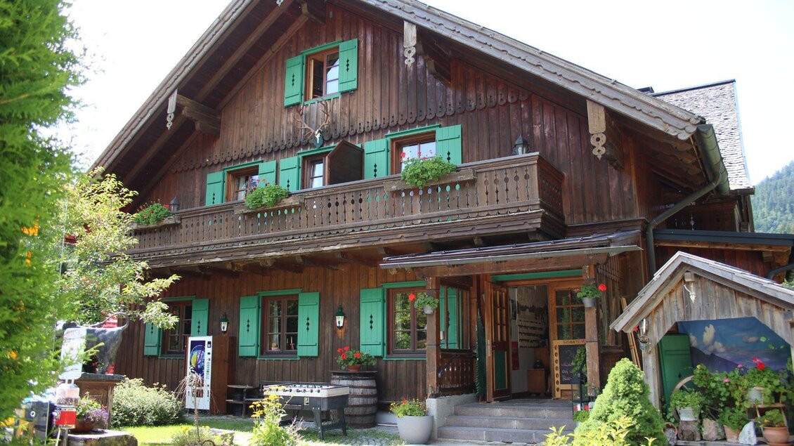 Ein traditionelles Holzhaus mit grünen Fenstern und einem Balkon. Der Garten ist gepflegt und mit Blumen dekoriert. | © Viola Lechner
