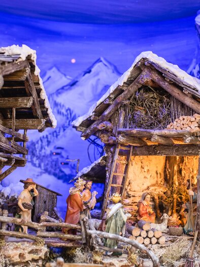 Eine detailreiche Krippe mit kleinen Figuren in einer winterlichen Landschaft. Im Hintergrund sind schneebedeckte Berge und zwei rustikale Holzhäuser zu sehen. | © photo-austria