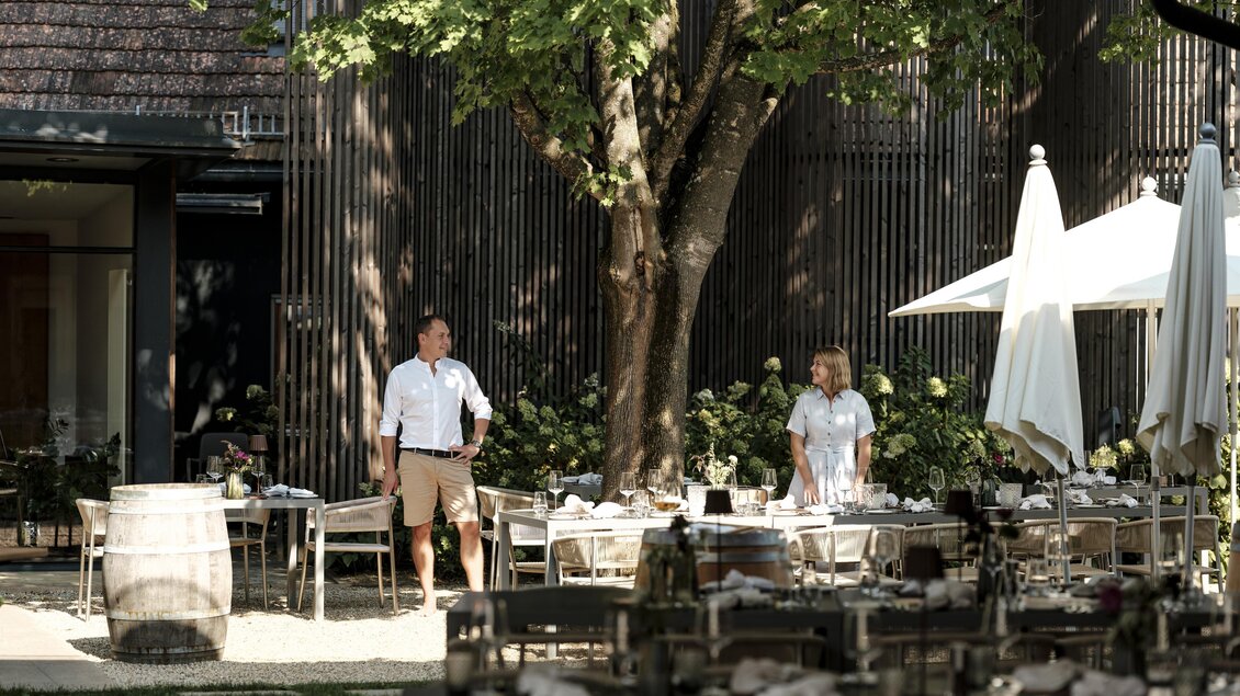 Ein einladendes Restaurant im Freien mit vielen Tischen und einem großen Baum. Zwei Personen stehen entspannt in der Umgebung. | © Reidinger