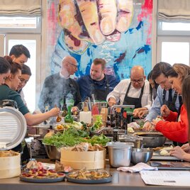 A group of people is preparing various dishes together in a modern kitchen. In the background, a colorful artwork can be seen. | © Rene Strasser