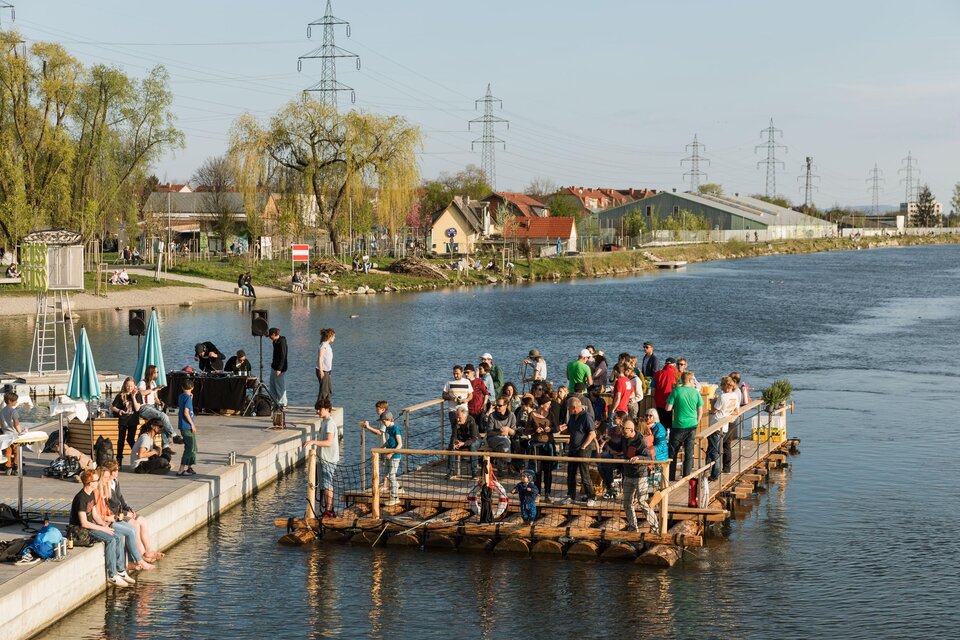 Eine Gruppe von Menschen steht auf einem Floß im Wasser, während andere am Ufer entspannen. Der Tag ist sonnig mit klarem Himmel und Blick auf die Umgebung. | © Die Flösserei