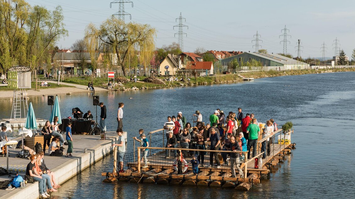 Eine Gruppe von Menschen steht auf einem Floß im Wasser, während andere am Ufer entspannen. Der Tag ist sonnig mit klarem Himmel und Blick auf die Umgebung. | © Die Flösserei
