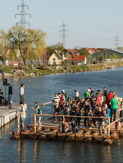 Eine Gruppe von Menschen steht auf einem Floß im Wasser, während andere am Ufer entspannen. Der Tag ist sonnig mit klarem Himmel und Blick auf die Umgebung. | © Die Flösserei